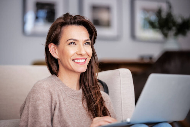 Close-up of an attractive middle aged woman sitting on armchair while using laptop. Beautiful female wearing casual clothes. Home office. - Foto, immagini