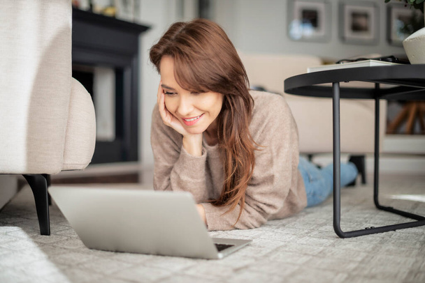 Full length shot of happy woman using laptop and having video call while relaxing at home. Attractive woman wearing sweater and blue jeans. - Fotó, kép