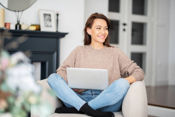 Shot of an attractive middle aged woman sitting on armchair while using laptop. Beautiful female wearing casual clothes.  - Foto, Bild