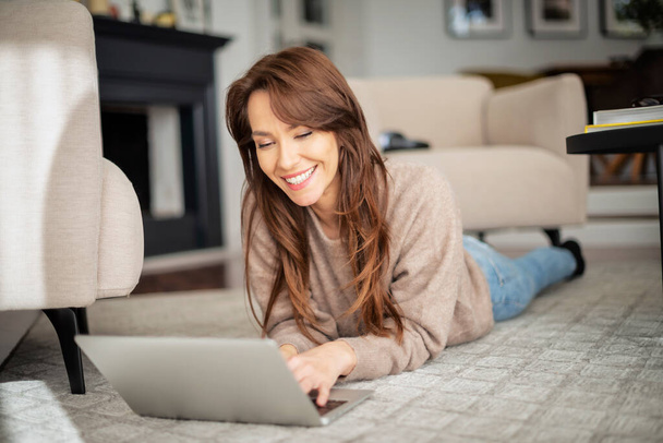 Full length shot of happy woman using laptop and browsing on the internet while relaxing at home. Attractive woman wearing sweater and blue jeans. - Zdjęcie, obraz