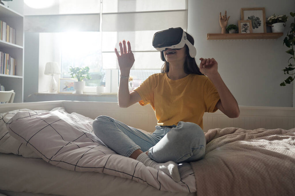 Adolescente donna caucasica utilizzando occhiali di realtà virtuale mentre seduto in camera da letto - Foto, immagini