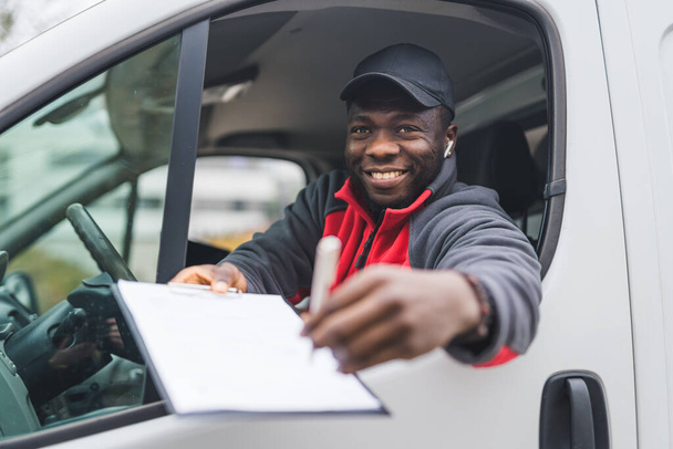 Young adult black delivery guy in red pullover sitting in white van smiling holding papers out to client to sget signed. Delivery service. Horizontal shot . High quality photo - Fotografie, Obrázek