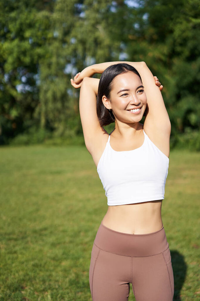 Asian woman stretching her arms, doing fitness workout in park, smiling pleased, warming up before jogging on fresh air in daytime. - Photo, Image
