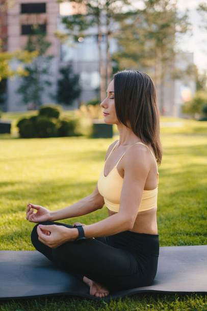 Porträt einer gesunden, reifen Frau, die frühmorgens meditiert und in Lotus-Pose auf einer Yogamatte in einem grünen Park sitzt. Meditation in der Natur - Foto, Bild
