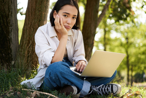 Nuoren aasialaisen naisen muotokuva istuu puistossa puun lähellä, työskentelee kannettavan tietokoneen kanssa ulkona. - Valokuva, kuva