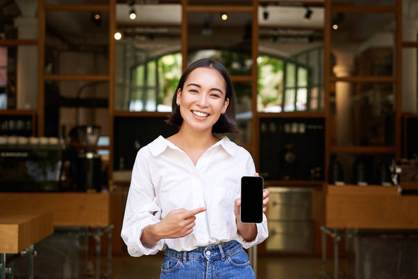 Happy asian woman, cafe manager standing in front of restaurant, pointing finger at smartphone app, showing mobile screen, recommending application. - Фото, зображення