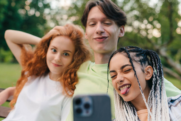 Fröhlich lächelnde Teenager entspannen sich an einem sonnigen Sommertag im Park und machen Selfies mit dem Handy. Modernes Fernkommunikationskonzept. Drei Personen. - Foto, Bild