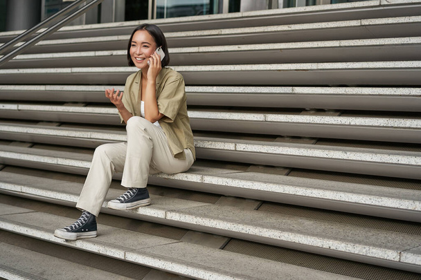 Улыбающаяся азиатка сидит на лестнице здания и разговаривает по мобильному телефону, расслабляясь во время телефонного разговора. - Фото, изображение