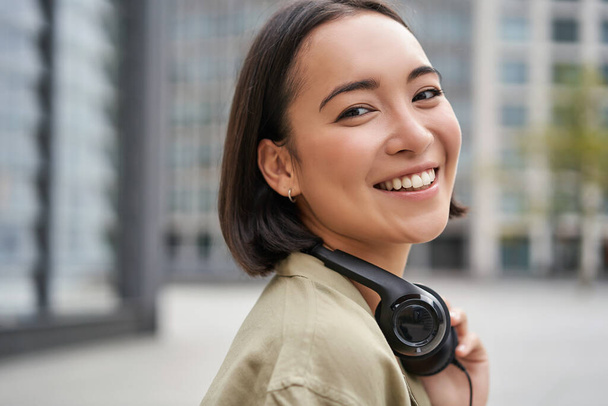 Portret szczęśliwej Azjatki w słuchawkach, lubi muzykę podczas spaceru po mieście, uśmiecha się i śmieje. - Zdjęcie, obraz