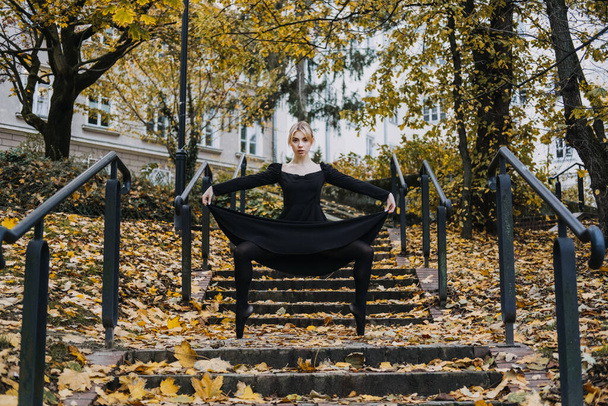 Ballerina Dancing in autumn city street, Modern Ballet Dancer in black dress, Pointe Shoes outdoors. Ballerina stretches, daily ballet stretching routine. - Foto, imagen