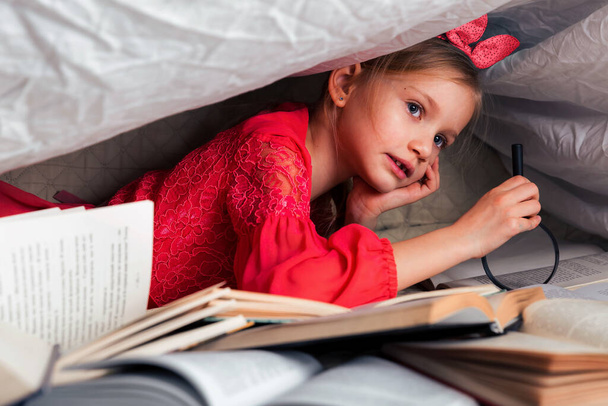 Cute 6-7 years old girl reads a book. Little girl at home on the bed in red dress under a blanket cover with a many books on foreground. Search information in book with magnifying glass - Fotó, kép