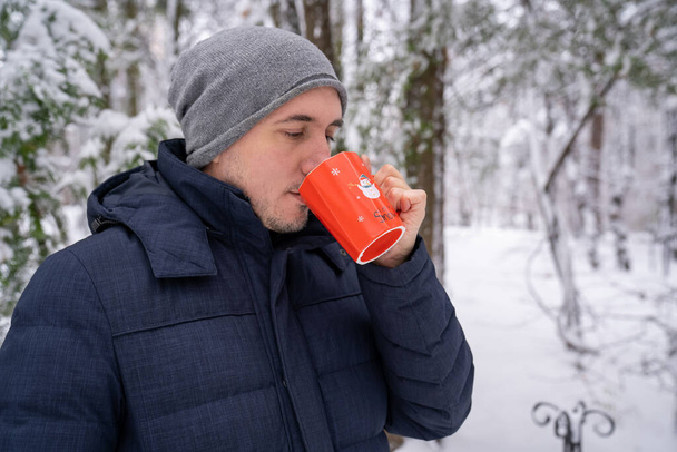 hombre guapo beber sabroso té caliente bebida de café al aire libre en el bosque de invierno nevado. La persona usa ropa casual sombrero gris y chaqueta de invierno. Cómo calentarse cuando hace frío.  - Foto, imagen