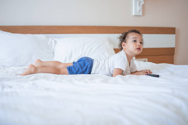 Küçük çocuk büyük yatak odasında beyaz bir yatakta karnının üstünde yatıyor. - Fotoğraf, Görsel