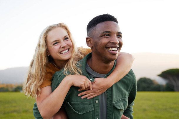 Happy multiracial young couple smiling and looking away during playful piggyback in field - 写真・画像