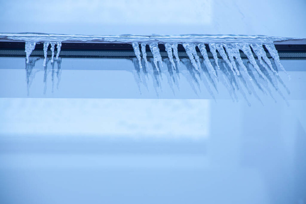 Παγοκρύσταλλοι κοντά στο παράθυρο το χειμώνα. Τζάμια πρόσοψης του κτιρίου με παγοκολώνες. Πάγος στα παράθυρα με κρύο καιρό. Μαλακή εστίαση στον παγοκρύσταλλο - Φωτογραφία, εικόνα