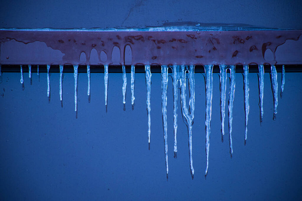 Παγοκρύσταλλοι κοντά στο παράθυρο το χειμώνα. Τζάμια πρόσοψης του κτιρίου με παγοκολώνες. Πάγος στα παράθυρα με κρύο καιρό. Μαλακή εστίαση στον παγοκρύσταλλο - Φωτογραφία, εικόνα