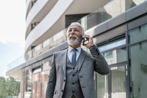 Homme d'affaires souriant parlant sur un téléphone intelligent. Professionnel masculin âgé confiant porte costume complet. Il est debout contre un immeuble de bureaux dans la ville. - Photo, image