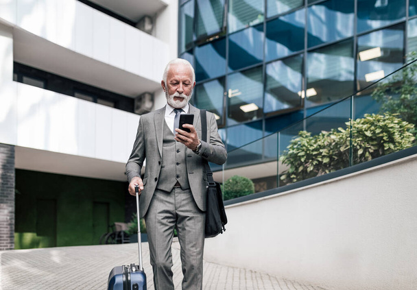 Starszy przedsiębiorca smsuje na smartfonie z torbą na laptopa. Starszy biznesmen w garniturze trzyma walizkę. Stoi przeciwko biurowcowi podczas podróży służbowych.. - Zdjęcie, obraz