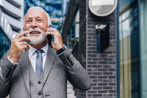 携帯電話で話す幸せな高齢男性の専門家。市内のオフィスビルに立っている間、自信を持っているシニア起業家がジェスチャーしている。彼はフルスーツを着ている. - 写真・画像