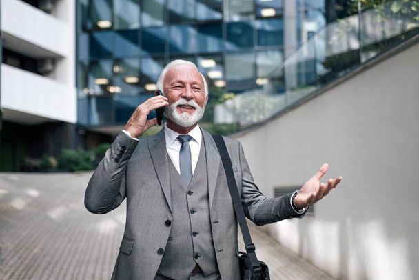 Onnellinen yrittäjä ele puhuessaan älypuhelimella. Hymyilevä vanhempi mies ammattilainen kannettavan tietokoneen laukku seisoo kaupungin business center. Hänellä on puku.. - Valokuva, kuva