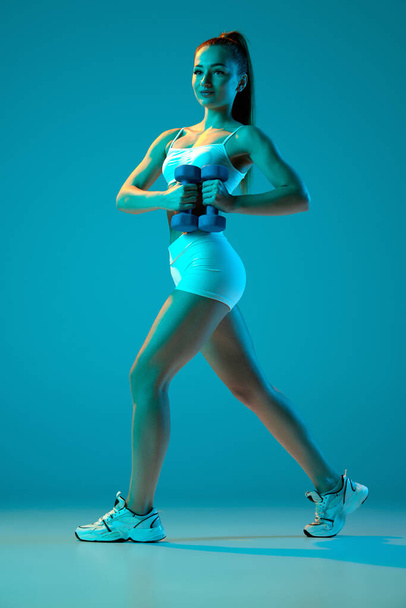 Young sportive girl training with dumbbells isolated over blue studio background in neon light. Strong hands. Modern sport, action, motion, youth concept. Fitness, hobby, healthy lifestyle - Фото, зображення