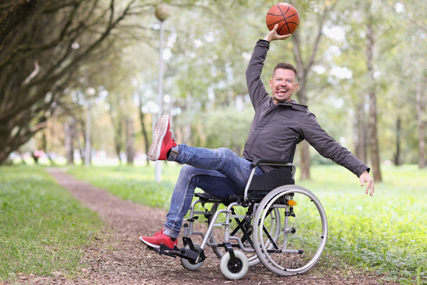 Portret van een grappige glimlachende gehandicapte man die met basketbal speelt. Een man in een rolstoel in een stadspark. Invaliditeits- en gezondheidsconcept - Foto, afbeelding