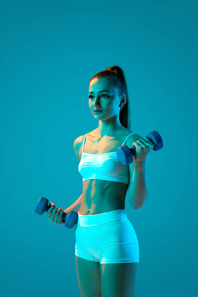 Young sportive girl in white sportswear training with dumbbells isolated on blue studio background in neon light. Muscular body. Modern sport, motion, youth concept. Fitness, hobby, healthy lifestyle - Photo, image