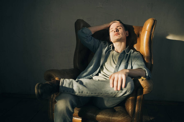Portret van Jonge stijlvolle man in denim of jeans kleding die op de fauteuil zit in de fotostudio. - Foto, afbeelding