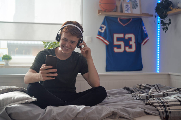 Καυκάσιος έφηβος αγόρι περιήγηση τηλέφωνο με χαμόγελο και φορώντας ακουστικά, ενώ κάθεται στο κρεβάτι - Φωτογραφία, εικόνα