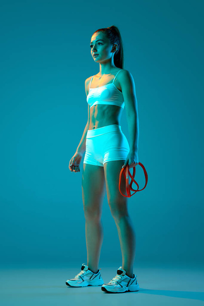 Young sportive girl posing with sports equipment, expanders isolated over blue studio background in neon light. Modern sport, action, motion, youth concept. Fitness, hobby, healthy lifestyle - Φωτογραφία, εικόνα