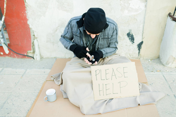 Arme dakloze man die de munten telt geld om voedsel te kopen terwijl hij op een karton zit met een teken van alsjeblieft helpen  - Foto, afbeelding