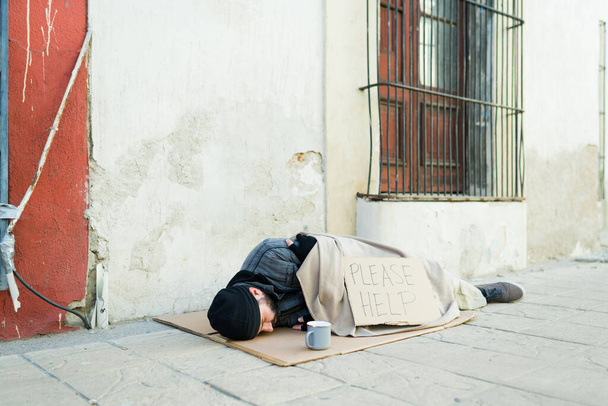 Homem sem-teto mendigo frio coberto com um cobertor dormindo na rua com um sinal de papelão por favor ajuda  - Foto, Imagem