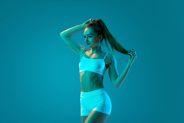 Young sportive girl in white sportswear posing isolated over blue studio background in neon light. Fit muscular body. Modern sport, action, motion, youth concept. Fitness, hobby, healthy lifestyle - Photo, image