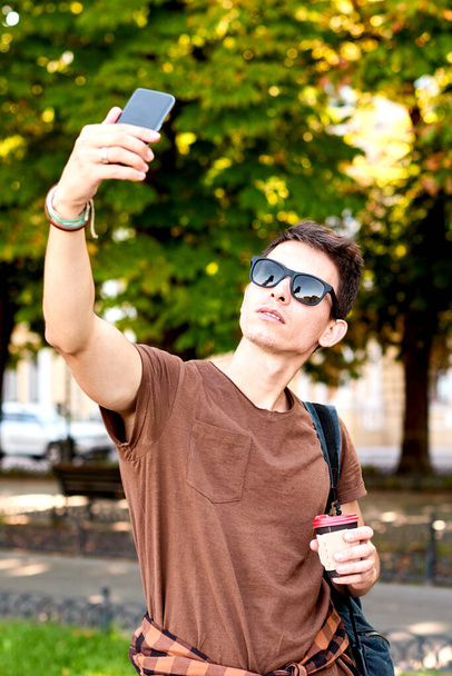 Jeune homme à la mode en T-shirt marron avec des lunettes de soleil prenant selfie dans la rue ou le parc de la ville - Photo, image