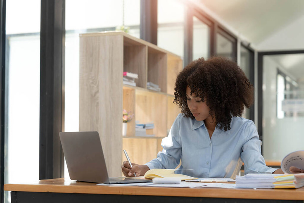 Joven empresaria africana sonriente sentada en un escritorio en su oficina en casa trabajando en línea con una computadora portátil
. - Foto, imagen