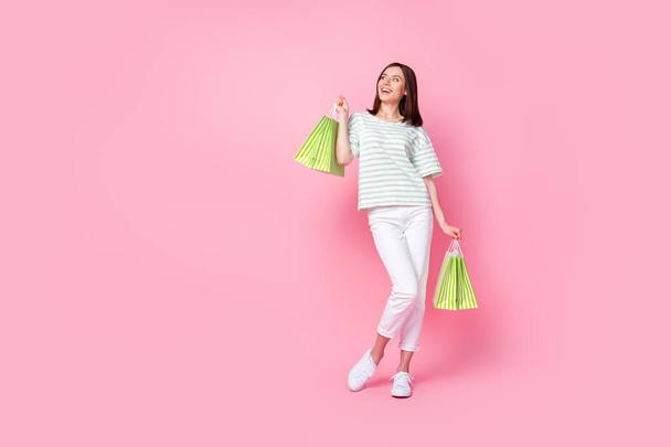 Foto de tamaño completo de elegante glamour chica vestida a rayas camiseta pantalones blancos zapatillas de deporte mirada espacio vacío aislado sobre fondo de color rosa. - Foto, imagen