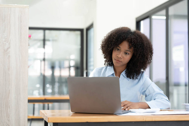 ノート パソコンでオンライン作業彼女のホーム オフィスの机に座って笑顔若いアフリカ女性起業家. - 写真・画像