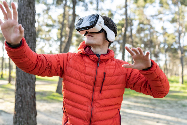 Καυκάσιος άνδρας με κόκκινο παλτό και γυαλιά εικονικής πραγματικότητας που παίζουν στο δάσος - Φωτογραφία, εικόνα