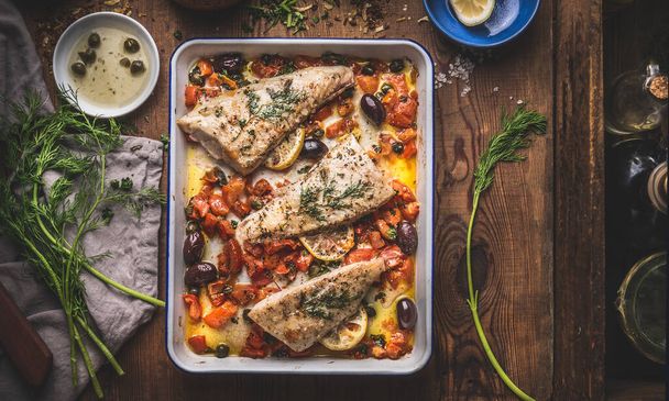 Tasty Bass fish fillets in Mediterranean sauce with tomatoes, olives and capers in baking pan on rustic wooden background with ingredients. Top view - Фото, зображення