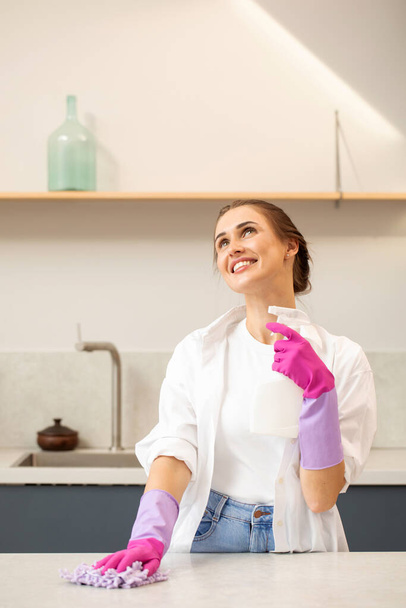 Een aantrekkelijke jonge vrouw in rubberen handschoenen houdt een fles wasmiddel vast en glimlacht het aanrecht in de keuken af. Het concept van schoonmaken van het huis. Schoonmaakmiddelen. - Foto, afbeelding