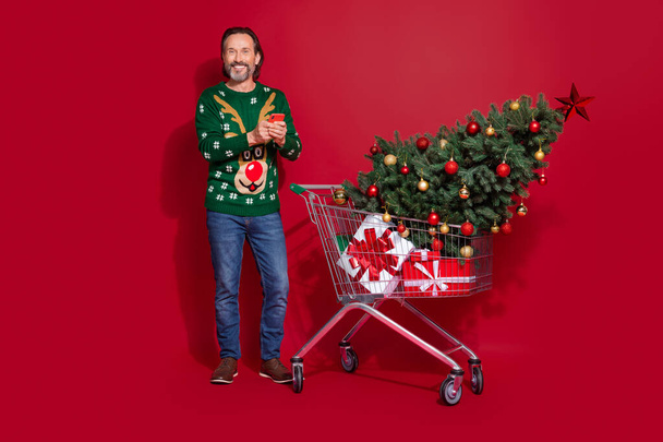 Full length portrait of positive handsome man hold use telephone hypermarket trolley evergreen giftbox isolated on red color background. - Fotoğraf, Görsel