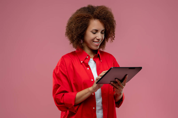 Na internetu. Hezká kudrnatá žena v červené bundě s tabletou v rukou - Fotografie, Obrázek