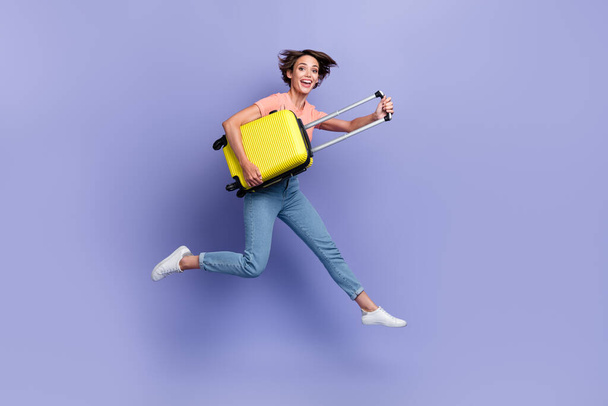 Full length photo of sweet excited girl dressed orange t-shirt jumping high holding suitcase isolated purple color background. - Foto, afbeelding