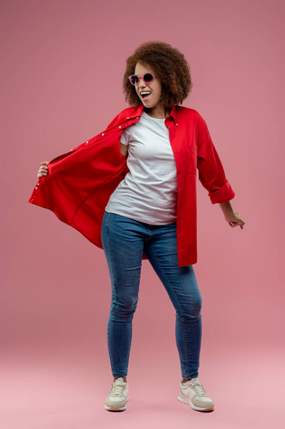 Model look. Pretty curly-haired woman in red jacket and sunglasses posing - Foto, Bild