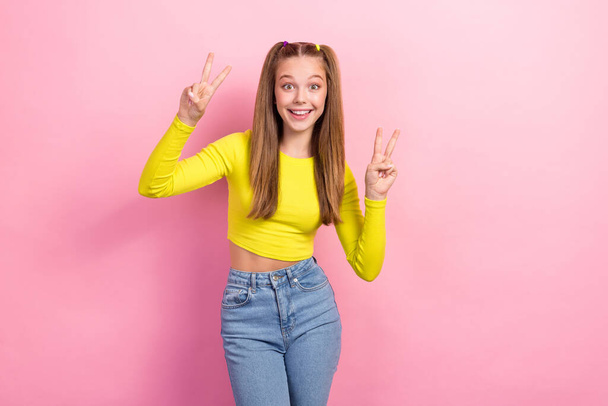 Photo of optimistic cute schoolgirl with ponytails dressed yellow long sleeve two hands showing v-sign isolated on yellow color background. - Fotografie, Obrázek