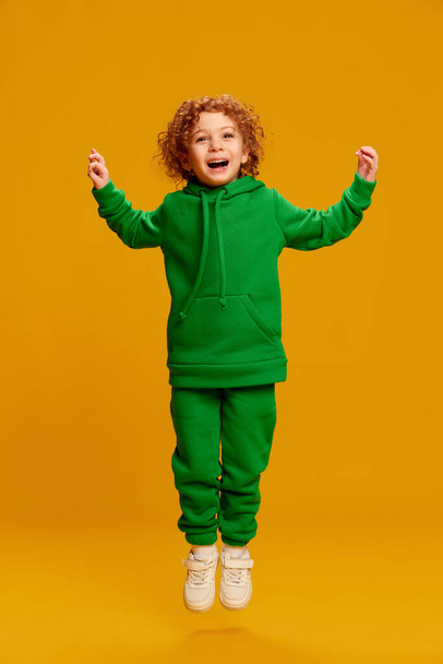 Portrait of cute little girl, child with curly red hair posing, jumping isolated over yellow background. Fun. Concept of childhood, emotions, lifestyle, fashion, happiness. Copy space for ad - Photo, image