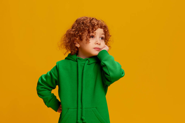 Portrait of cute little girl, child with curly red hair posing with bored face isolated over yellow background. Concept of childhood, emotions, lifestyle, fashion, happiness. Copy space for ad - Foto, Imagen