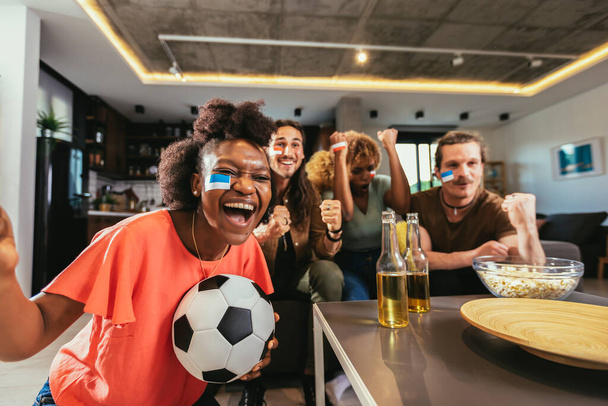 Young group watching sports on television and cheering. Emotional football fans watching football game together at home. Sports Fans with Painted Faces Sitting on a Couch Watch Game on TV - 写真・画像
