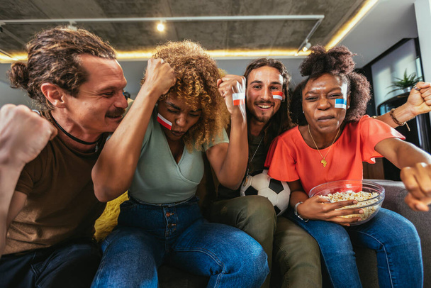 Young group watching sports on television and cheering. Emotional football fans watching football game together at home. Sports Fans with Painted Faces Sitting on a Couch Watch Game on TV - Foto, afbeelding