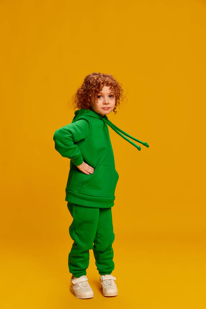 Portrait of cute little girl, child with curly red hair posing in green costume isolated over yellow background. Concept of childhood, emotions, lifestyle, fashion, happiness. Copy space for ad - Photo, image